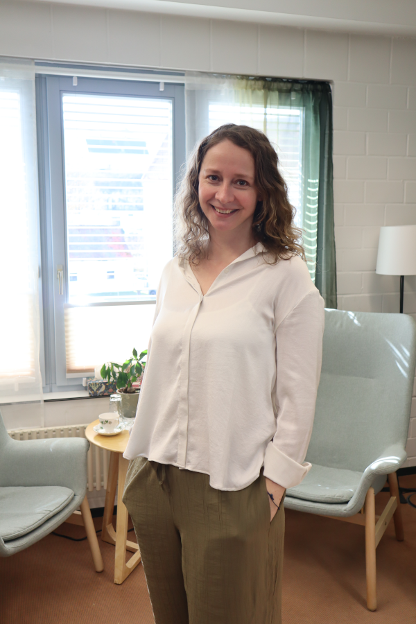 Diplom Psychologin Daniela Schötz-Mellor in ihrer Psychologischen Praxis in Sinzing in der Nähe Regensburg. Sie steht in einer lockeren Haltung, die Hände in den Hosentaschen. Gekleidet mit einer weißen Bluse und einer grünen Hose. Im Hintergrund sind helle Stühle zu sehen